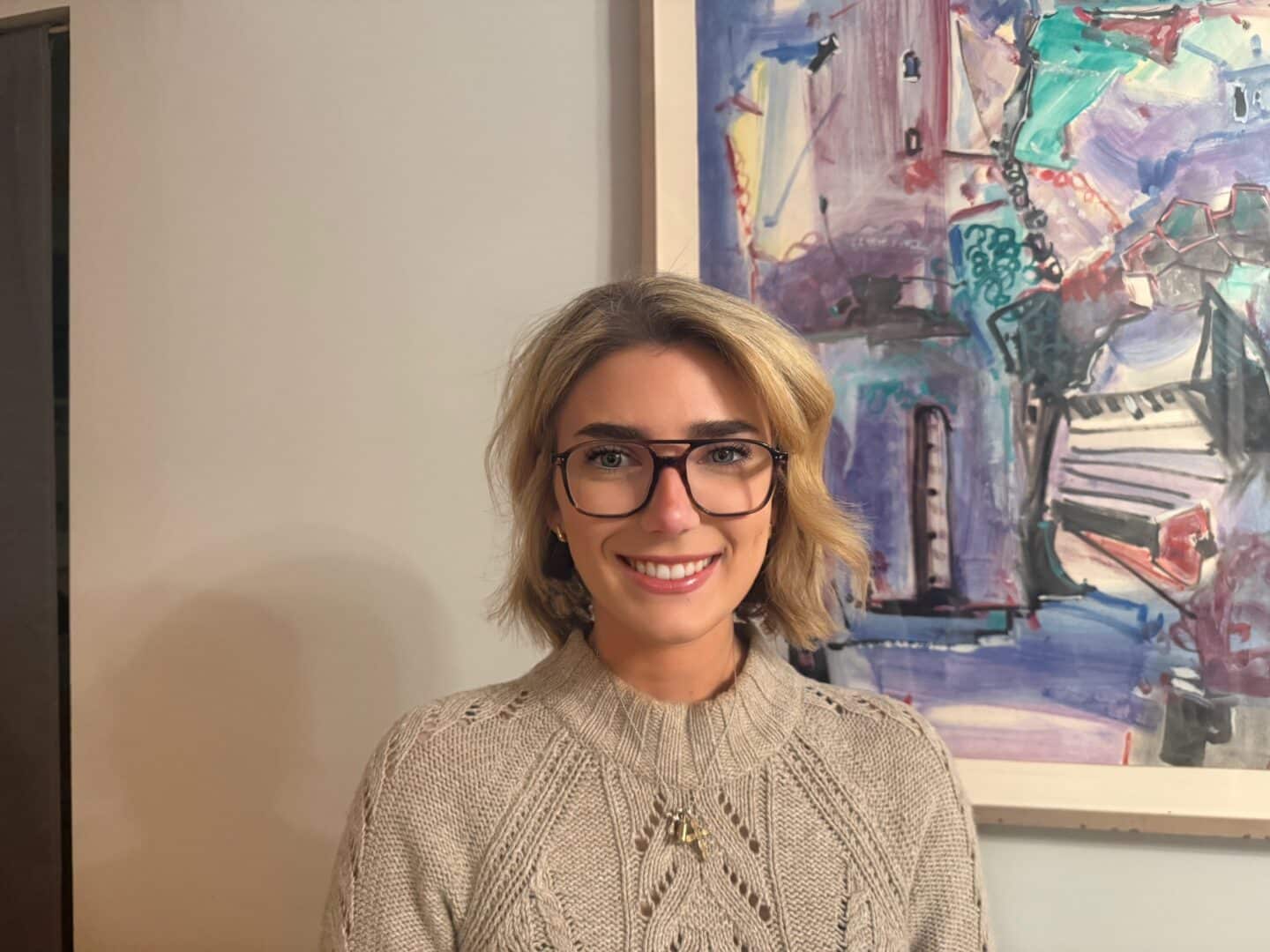 A smiling person with short blonde hair wearing glasses and a light-colored knit sweater, standing indoors in front of an abstract painting on the wall.