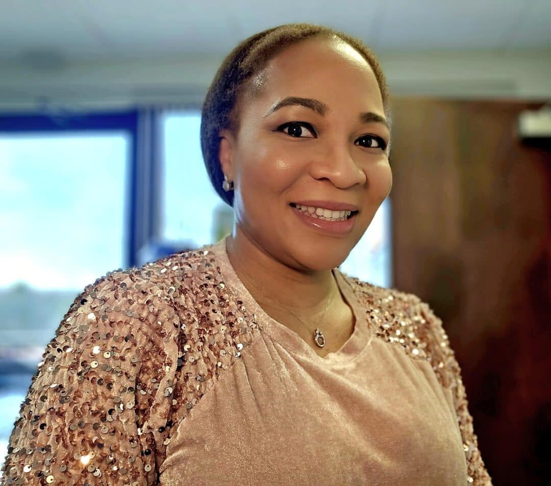A smiling person wearing a light pink, sequined top and a delicate necklace, standing indoors near a window with soft natural light.
