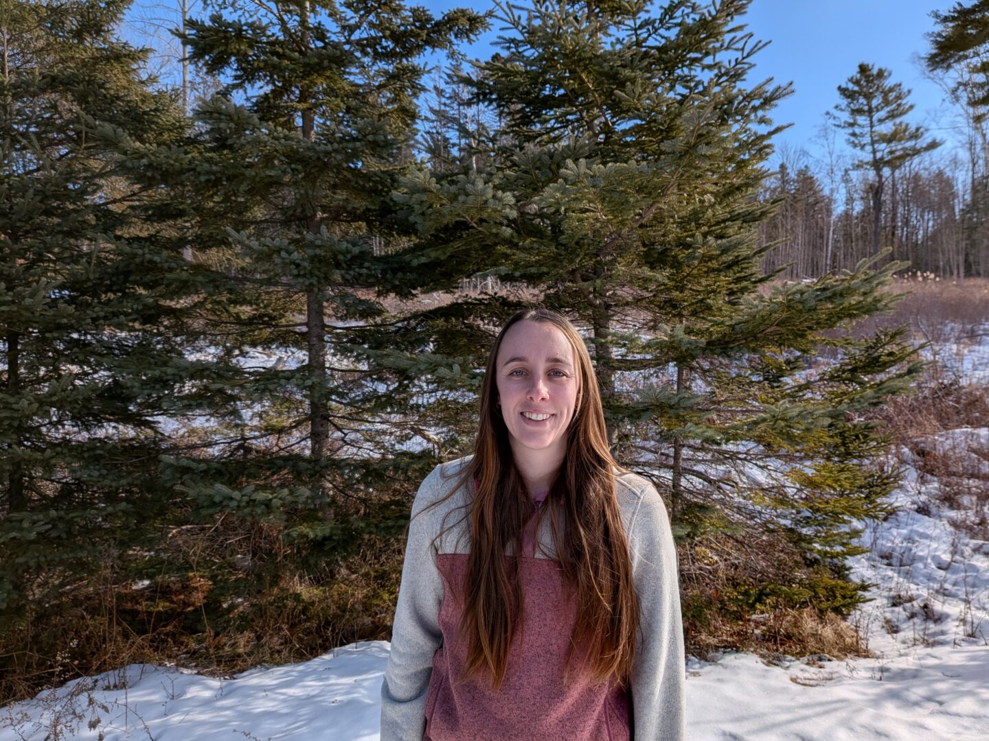 A smiling person with long brown hair wearing a two-tone sweater, standing outdoors in front of evergreen trees with patches of snow on the ground under a clear blue sky.